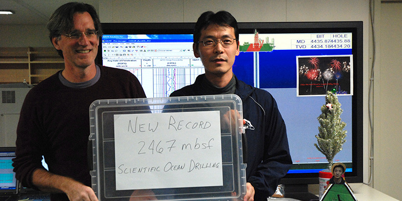 Photo of IODP Exp 348 co-Chief Scientists, Drs. Harold Tobin (L) and Takehiro Hirose (R) showing the new depth record achieved by <i>Chikyu</i> in December in 2013, 2,467 m below sea floor. This was beaten in January 2014, when <i>Chikyu</i> increased the borehole to 3058.5 m by the end of the expedition.